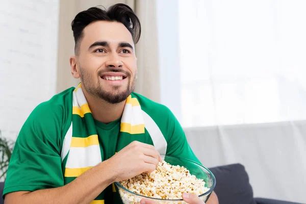 Cheerful sports fan with bowl of popcorn watching game at home — Photo de stock