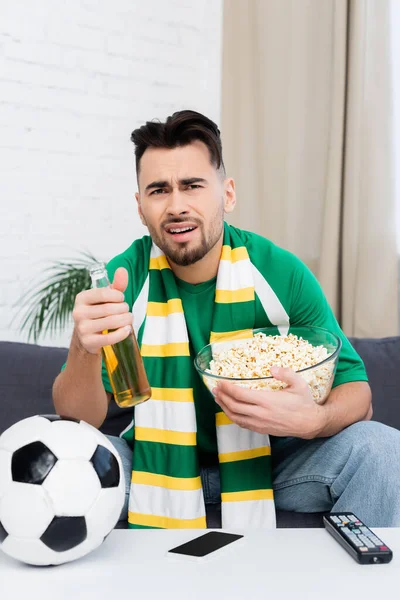 Displeased sports fan with popcorn and beer watching game on tv near soccer ball — Stockfoto