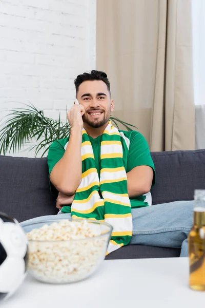 Fã de futebol feliz falando no smartphone perto de pipocas borradas e cerveja — Fotografia de Stock