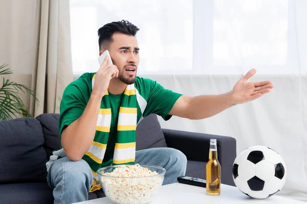 Ventilador de esportes descontente apontando com a mão e falando no smartphone enquanto assiste jogo de futebol na tv — Fotografia de Stock