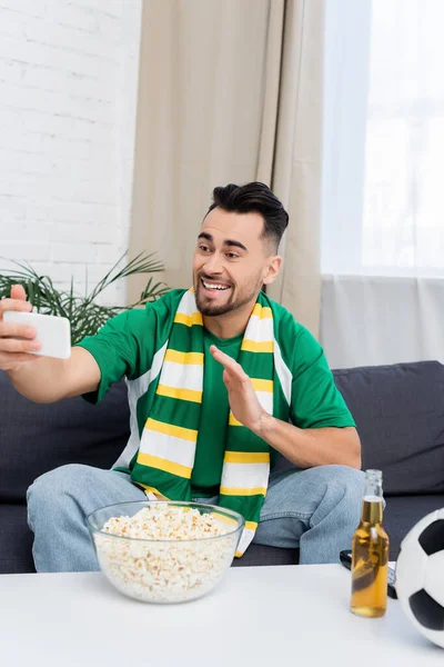 Cheerful sports fan waving hand during video call near popcorn and beer — Stockfoto