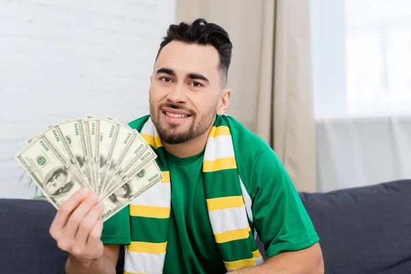 Man in scarf of sports fan looking at camera and showing dollar banknotes — Stock Photo