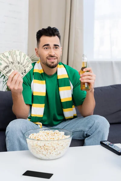 Disappointed sports fan with money and beer watching match at home — Stock Photo