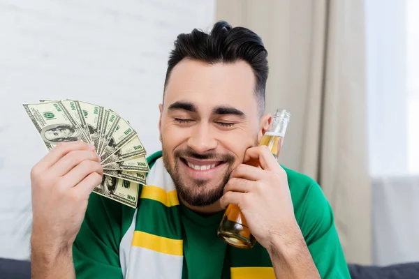 Pleased bookmaker with closed eyes holding dollar banknotes and beer — Stock Photo