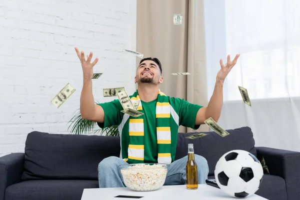 Happy bookmaker throwing up dollar banknotes near soccer ball, beer and popcorn — Photo de stock