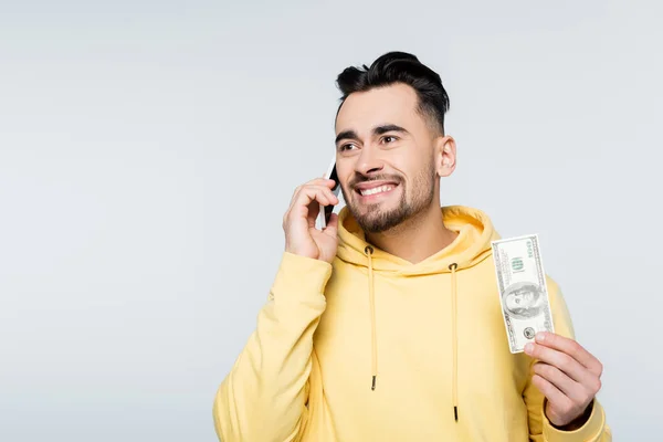 Joyful bookmaker with dollar banknote talking on smartphone isolated on grey — стоковое фото