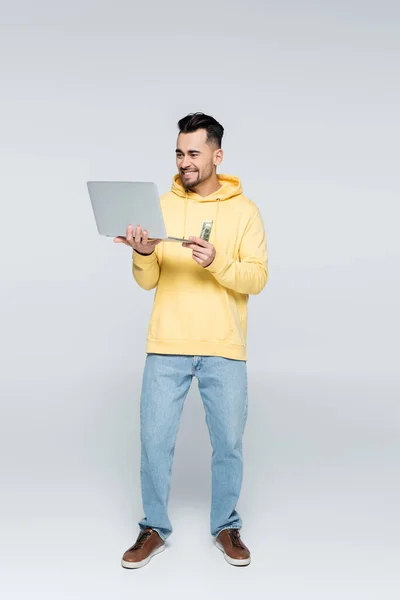 Full length view of happy gambler with dollars and laptop on grey - foto de stock