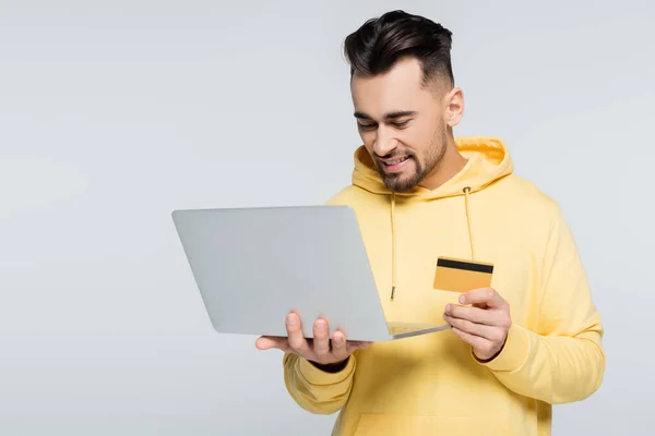 Uomo soddisfatto in felpa gialla con cappuccio in possesso di carta di credito vicino al computer portatile isolato su grigio — Foto stock