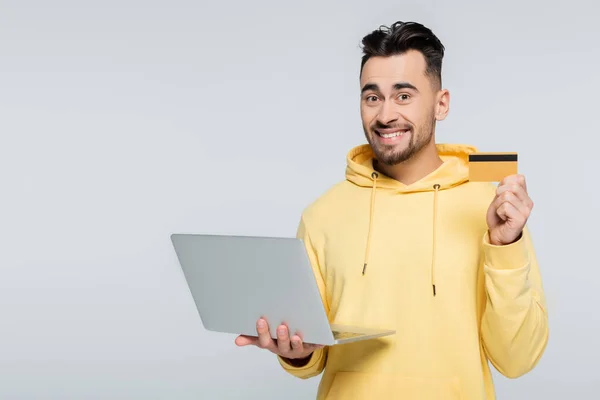 Homem satisfeito segurando cartão de crédito e laptop enquanto olha para a câmera isolada no cinza — Fotografia de Stock