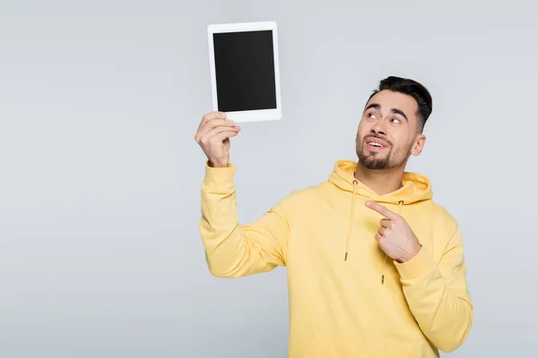 Uomo positivo in felpa gialla con cappuccio che punta al tablet digitale con schermo bianco isolato su grigio — Foto stock