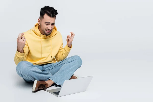 Worried gambler showing crossed fingers while sitting with laptop on grey — Stock Photo
