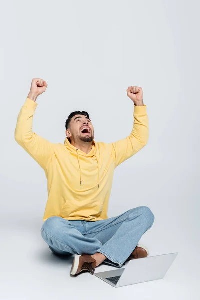 Jugador emocionado sentado cerca de la computadora portátil con las piernas cruzadas y mostrando el gesto de triunfo en gris - foto de stock