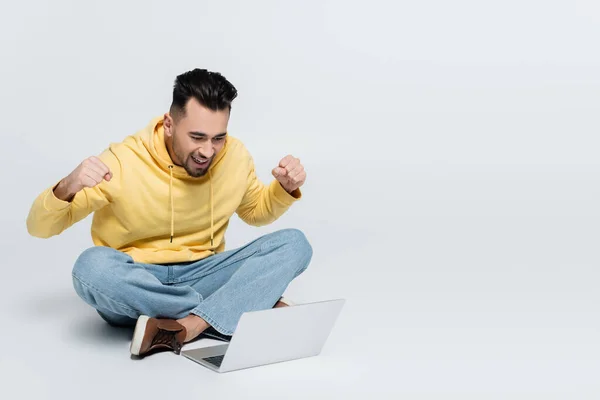 Cheerful man showing triumph gesture while sitting with laptop on grey — Stockfoto