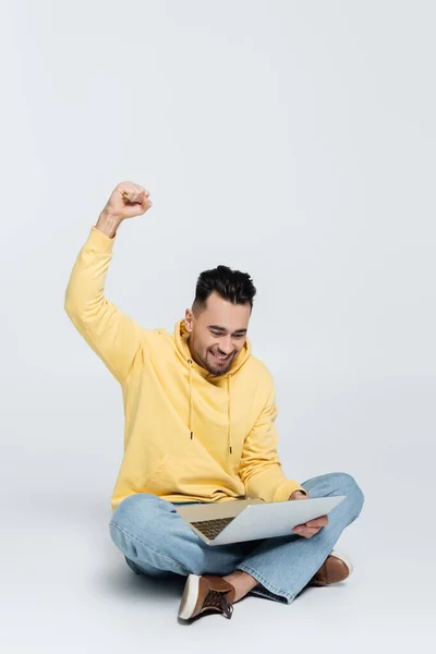 Excited bookmaker showing win gesture while sitting with laptop on grey - foto de stock