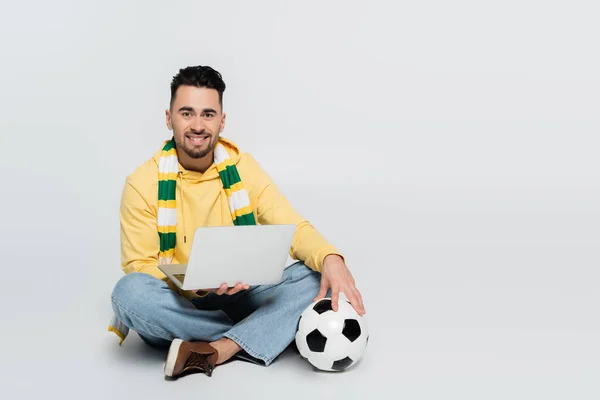 Jugador satisfecho sentado con el ordenador portátil y pelota de fútbol mientras mira a la cámara en gris - foto de stock