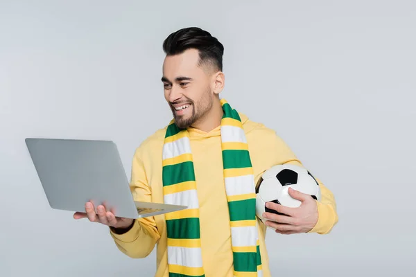 Corredor de apuestas feliz con pelota de fútbol mirando portátil aislado en gris - foto de stock