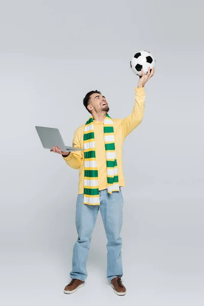 Comprimento total da casa de apostas com laptop segurando bola de futebol na mão levantada no cinza — Fotografia de Stock