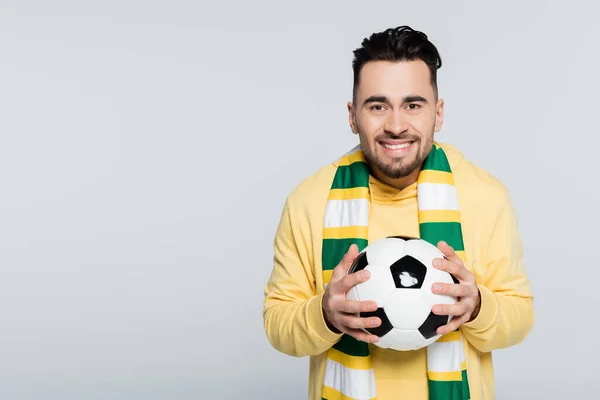 Веселий чоловік в смугастому шарфі тримає футбольний м'яч і дивиться на камеру ізольовано на сірому — стокове фото