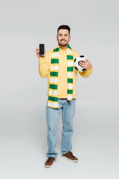 Full length of happy gambler with soccer ball showing cellphone with blank screen on grey — стоковое фото