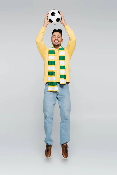 Full length of overjoyed sports fan jumping with soccer ball in raised hands on grey — Stockfoto