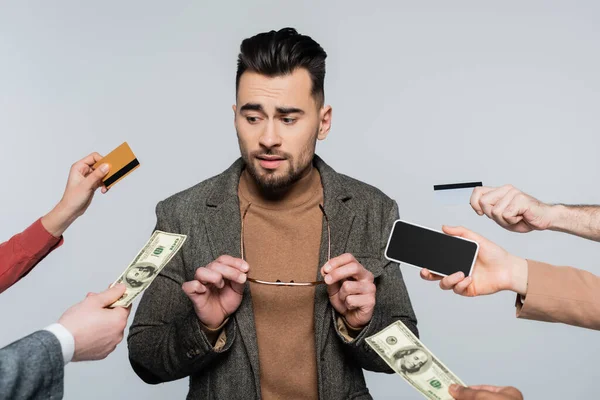 Jovem crítico segurando óculos perto de pessoas com dinheiro, cartões de crédito e smartphone isolado em cinza — Fotografia de Stock