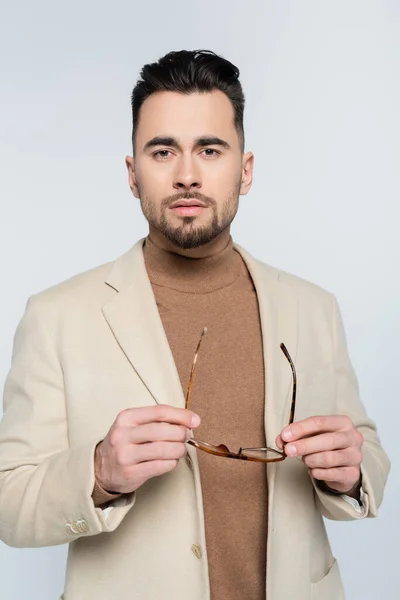 Young critic in beige blazer holding eyeglasses and looking at camera isolated on grey — Stock Photo