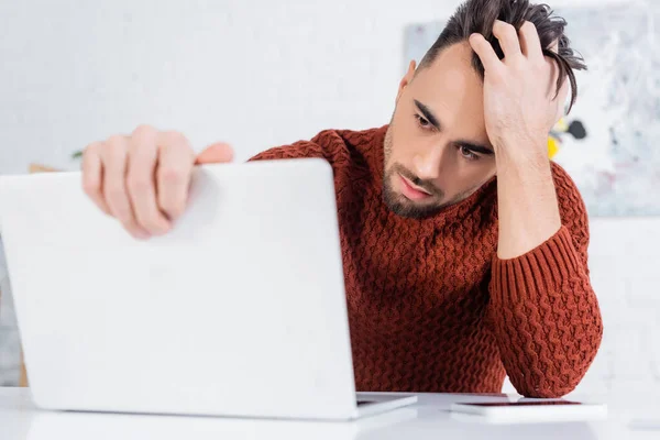 Depressed bookmaker touching head while looking at blurred laptop - foto de stock