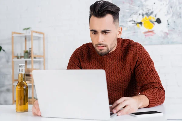 Focused gambler looking at laptop near bottle of beer and smartphone — Stock Photo