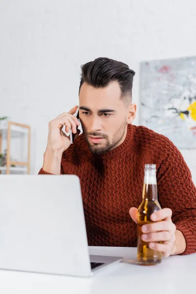 Serious bookmaker with bottle of beer talking on smartphone near laptop — Stock Photo