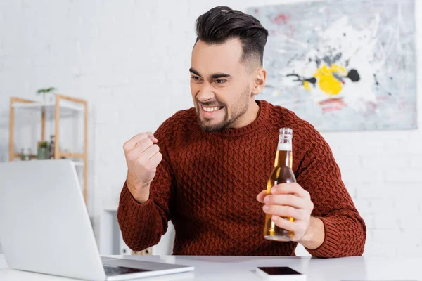 Захоплюючий гравець з пляшкою пива, що показує жест виграшу біля ноутбука — стокове фото