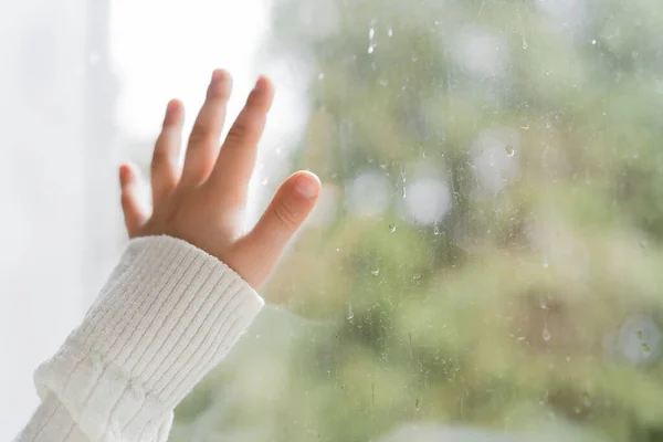 Vista recortada de la mano del niño cerca de la ventana con gotas de lluvia - foto de stock