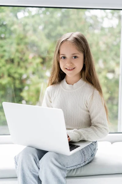 Chica feliz mirando a la cámara mientras está sentado cerca de la ventana con el ordenador portátil - foto de stock