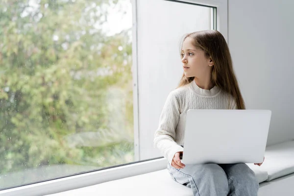 Menina olhando para longe enquanto sentado no peitoril da janela com laptop — Fotografia de Stock