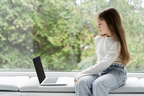 Menina sentada no peitoril da janela em casa perto do laptop com tela em branco — Fotografia de Stock