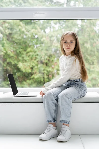 Ganzkörperansicht des Mädchens, das auf der Fensterbank neben dem Laptop sitzt und in die Kamera schaut — Stockfoto