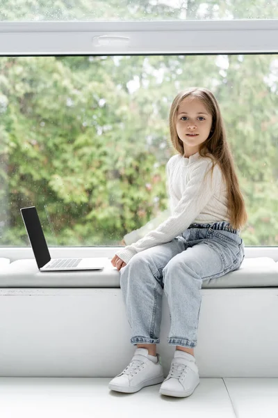 Full length view of girl looking at camera while sitting on windowsill near laptop — Stock Photo