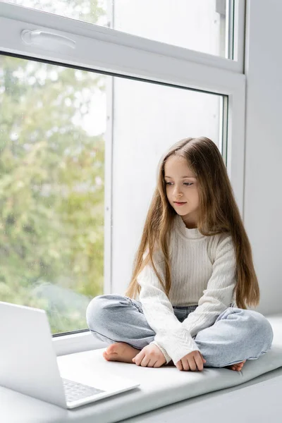 Chica descalza con el pelo largo sentado en el alféizar de la ventana cerca de la computadora portátil - foto de stock