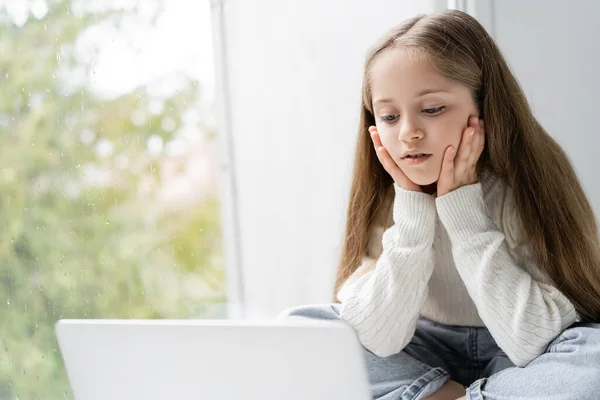 Focused girl holding hands near face while looking at laptop near window — Stock Photo