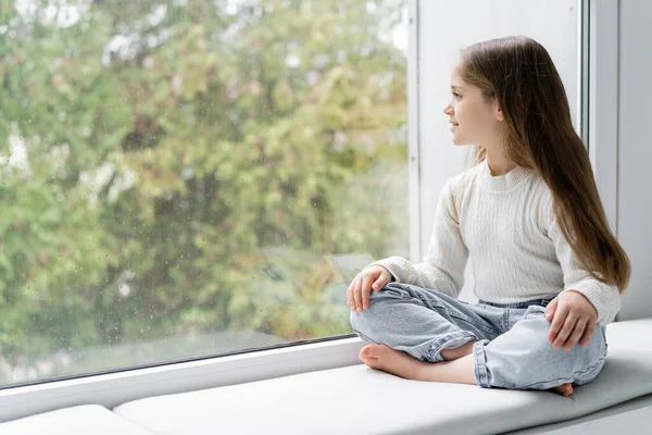 Chica descalza en jeans sentada en el alféizar de la ventana con las piernas cruzadas y mirando a través de cristal con gotas de lluvia - foto de stock