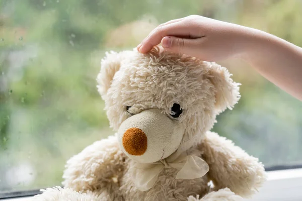 Abgeschnittene Ansicht von Kind berührt Teddybär in der Nähe verschwommenes Fenster mit Regentropfen — Stockfoto