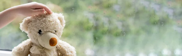 Vista parcial de la mano del niño cerca del oso de peluche y ventana borrosa con gotas de lluvia, pancarta - foto de stock