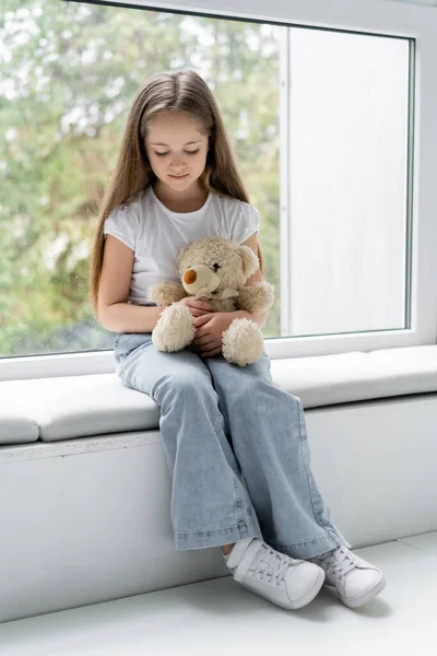 Vista de longitud completa de la niña sosteniendo oso de peluche mientras está sentado en el alféizar de la ventana - foto de stock