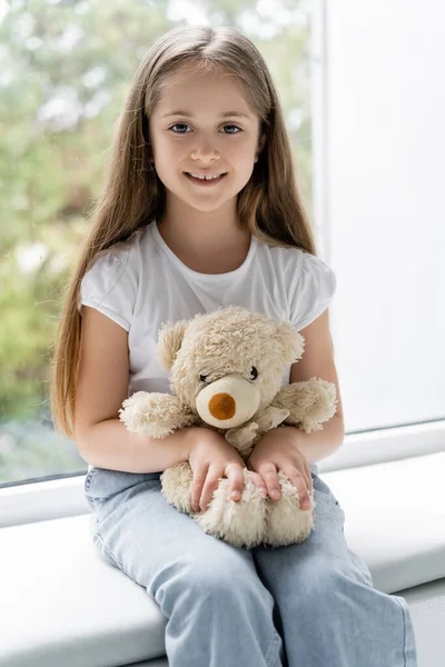 Cheerful girl smiling at camera while sitting near window with teddy bear — Stock Photo