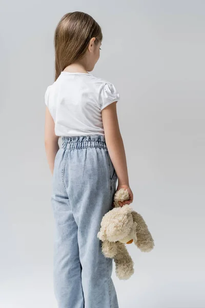 Вид ззаду розчарованої дівчини, що стоїть з плюшевим ведмедем ізольовано на сірому — стокове фото