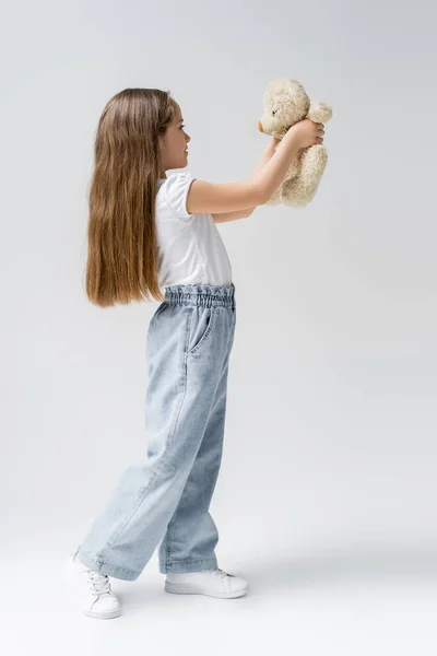 Comprimento total da menina em jeans segurando ursinho de pelúcia em cinza — Fotografia de Stock