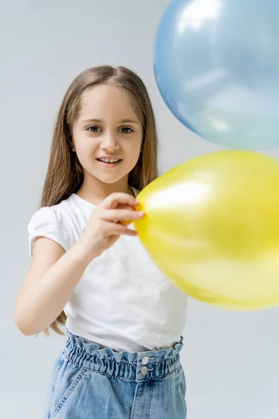 Chica complacida mirando a la cámara cerca borrosa globos azules y amarillos aislados en gris - foto de stock
