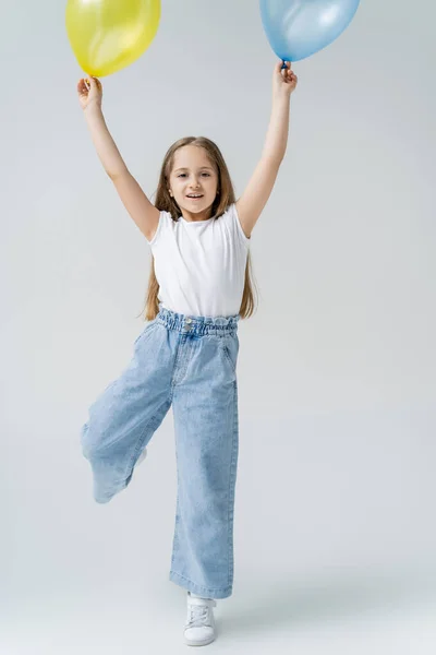 Full length view of cheerful girl in jeans holding blue and yellow balloons in raised hands on grey — Stock Photo