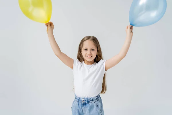 Happy girl in white t-shirt standing with blue and yellow balloons in raised hands isolated on grey — Stock Photo