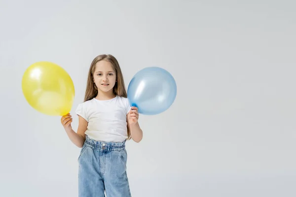 Glückliches Mädchen mit blauen und gelben Luftballons, das isoliert auf grau in die Kamera blickt — Stockfoto