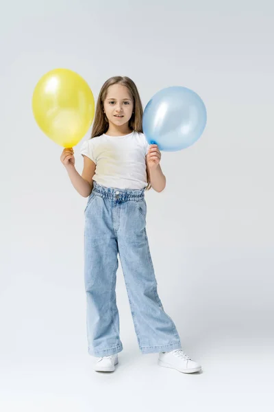 Piena vista lunghezza della ragazza ucraina in jeans in piedi con palloncini blu e gialli su grigio — Foto stock
